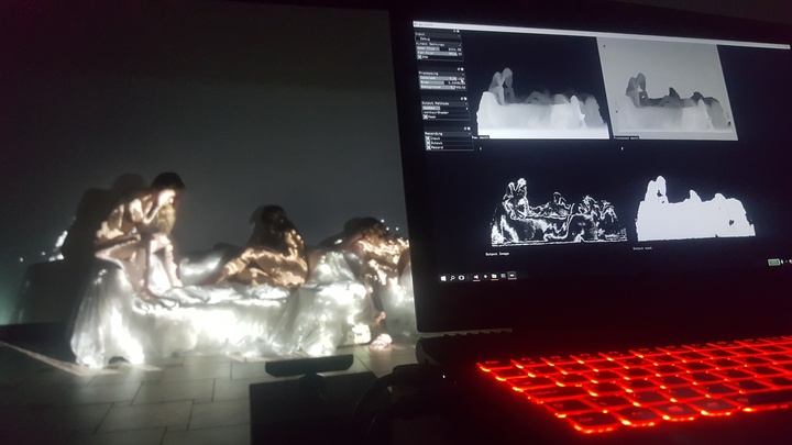Performers draped in gold-lit fabric sit on a lit platform beside a monitor showing depth-camera silhouette processing.