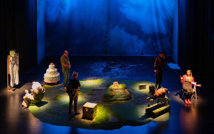 Stage set with a tiered cake, carousel horse and giant die on grassy mounds, performers in headphones.