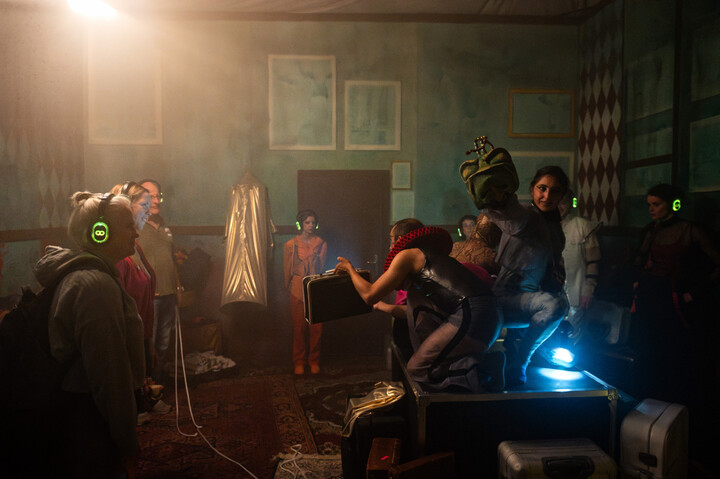 Performers in costume, including a frog-masked figure, greet audience in a smoky backstage room.
