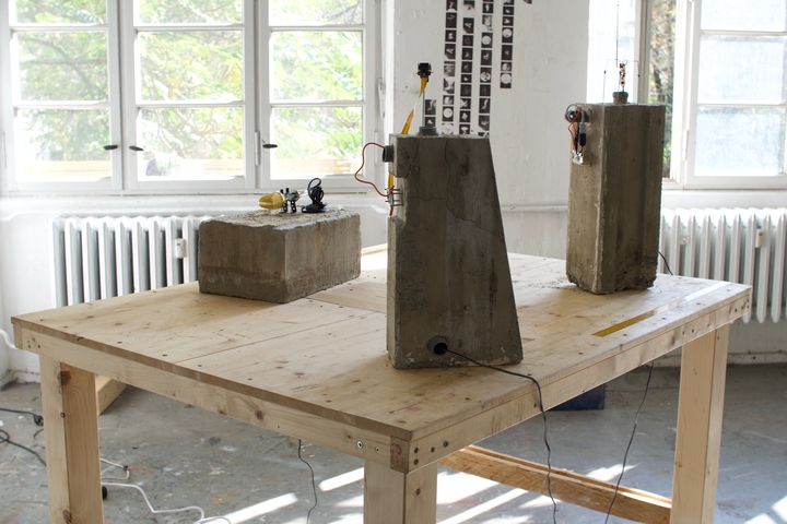 Raw concrete sculptures fitted with cameras and circuit boards stand on a wooden workbench in a bright studio.