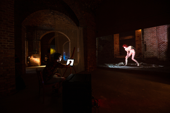 In a brick vault, a visitor draws on a tablet while a nude figure is projected life-size onto the far wall.