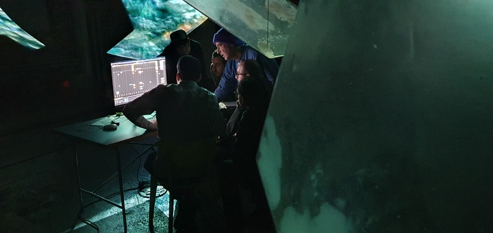 A small group gathers around a monitor showing music sequencer software during a dim rehearsal for Oumuamua.