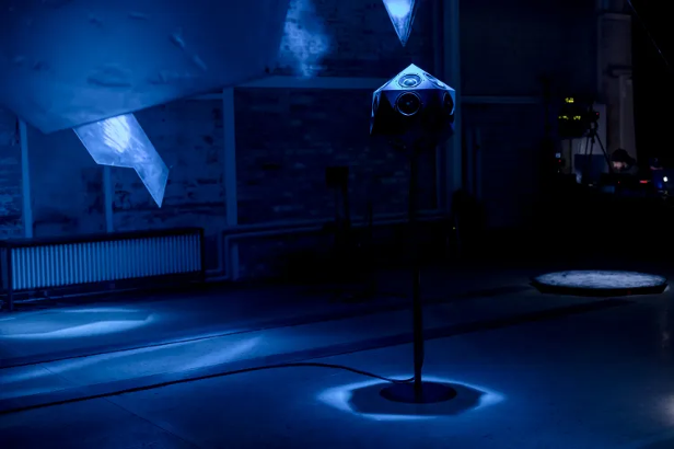 A faceted speaker sculpture on a stand glows blue in a dark installation space for Oumuamua.