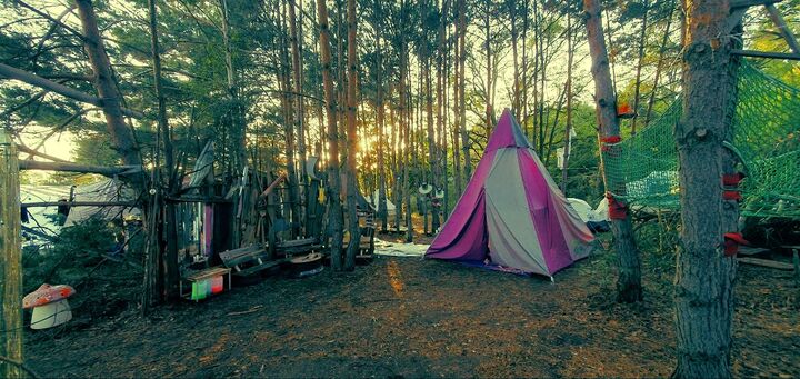 Purple-and-white teepee pitched among pine trees at a Burning Man-style camp, with a mushroom sculpture nearby at sunset.