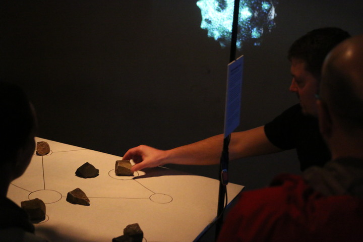 A visitor places a stone onto a circle in a diagram drawn on paper, with a glowing blue projection visible behind them.