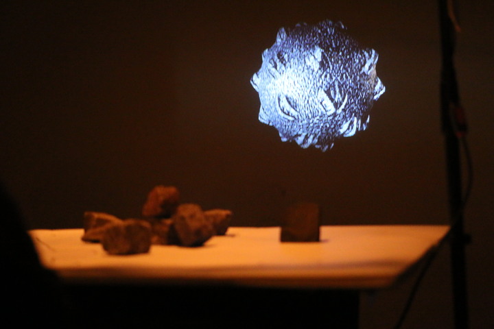 A cluster of stones sits on a table beneath a floating, spiky blue-lit projected form.
